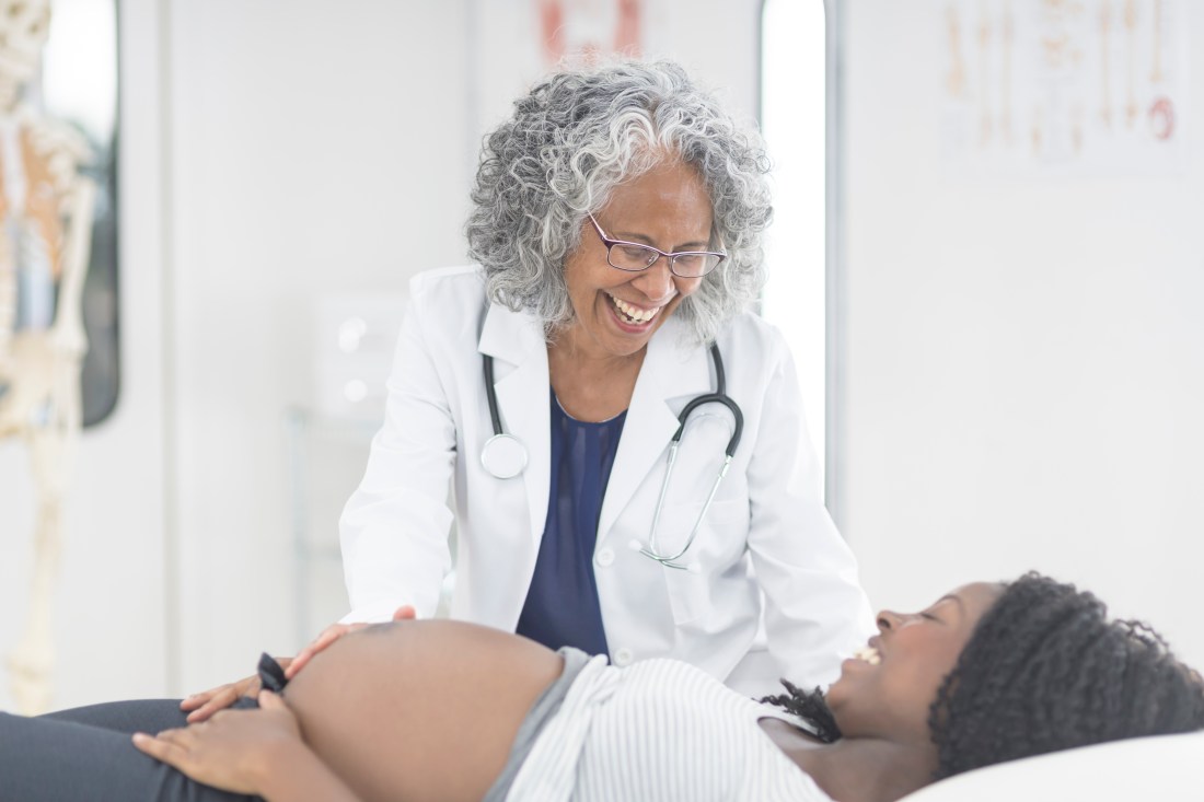 A pregnant woman is laying down on an examination table at the doctor's office. Her shirt is pulled up so you can see her pregnant belly. The female physician and patient are smiling and looking at one another.